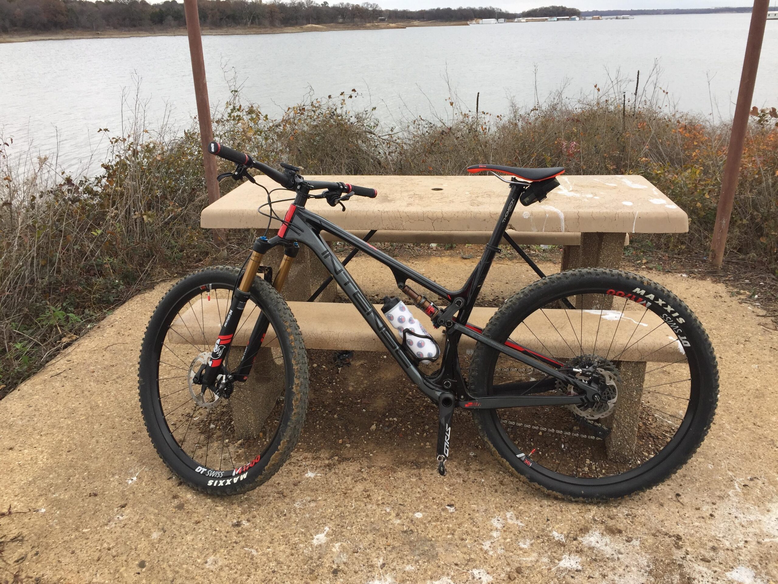 Intense Sniper Trail: A mountain bike with a black frame and red accents is parked next to a concrete picnic table by a lake. The surrounding area features sparse vegetation and a serene water view, with a cloudy sky above.