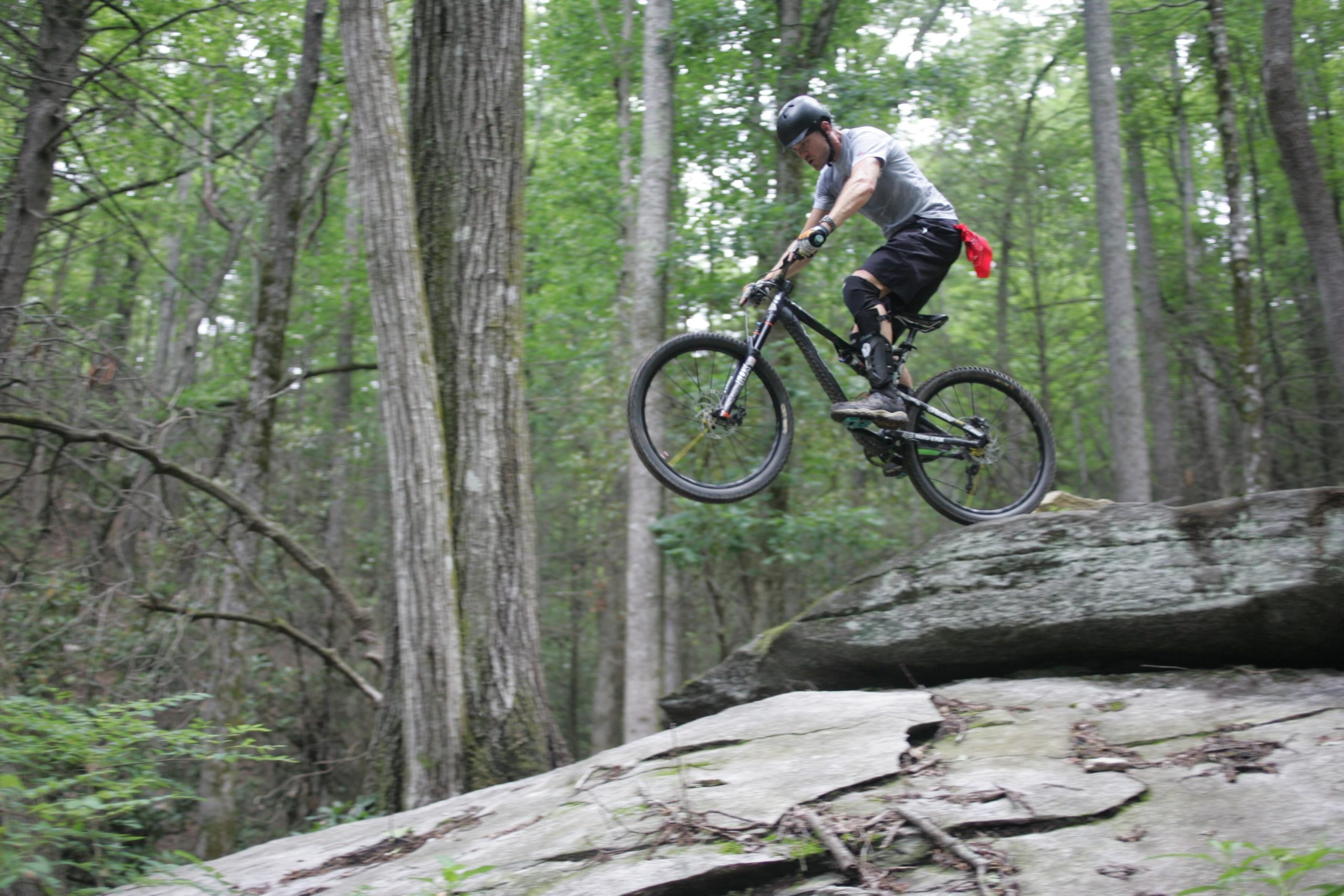A mountain biker wearing protective gear performs a jump off a large rock in a green forest. The biker is mid-air with the bike positioned vertically, surrounded by tall trees and foliage. Weed Patch Mountain Trail mountain bike trail.