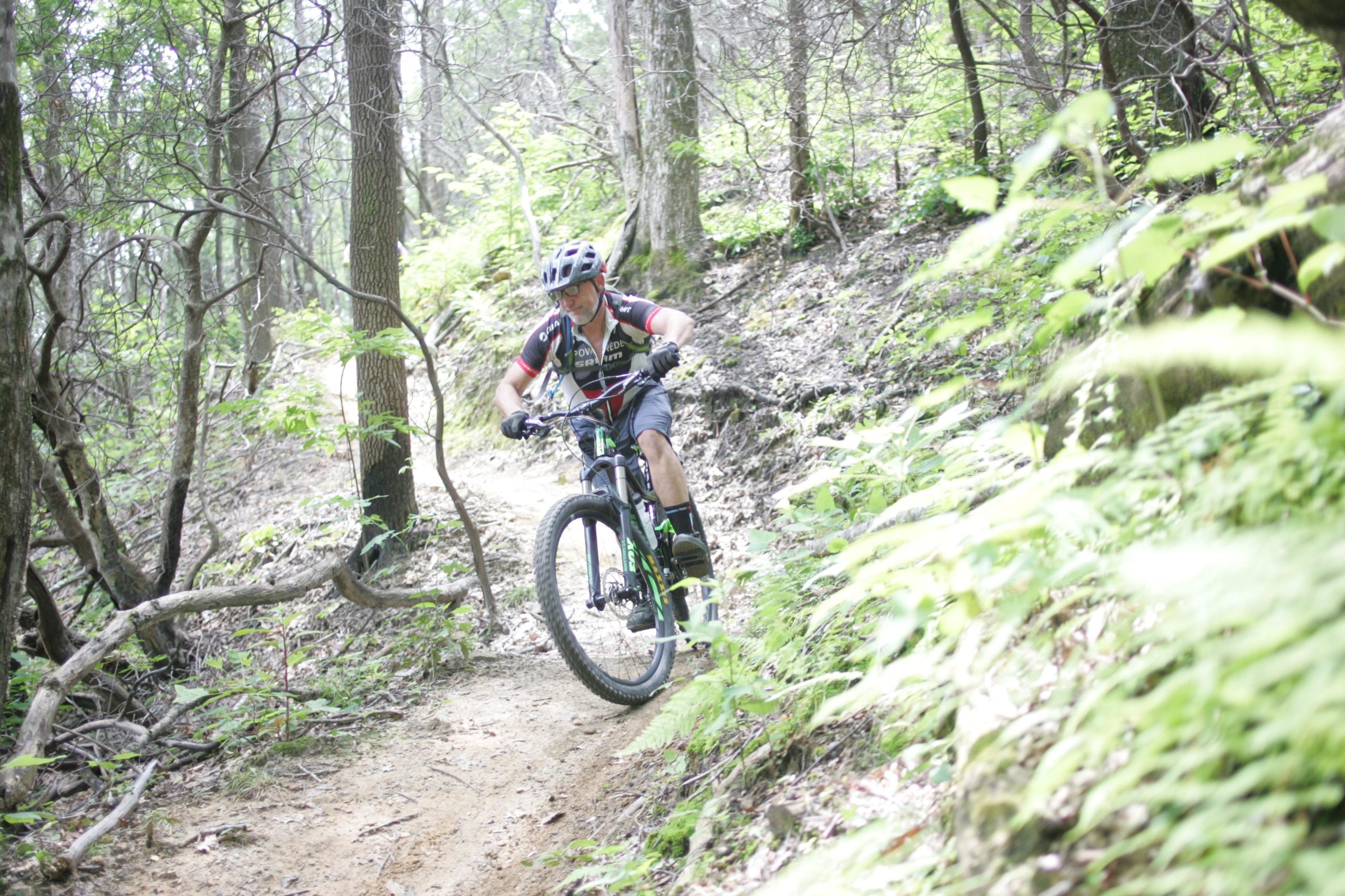 A person riding a mountain bike on a narrow dirt trail surrounded by green foliage and trees in a forested area. The cyclist is leaning forward, navigating the terrain, and wearing a helmet and biking gear. Weed Patch Mountain Trail mountain bike trail.