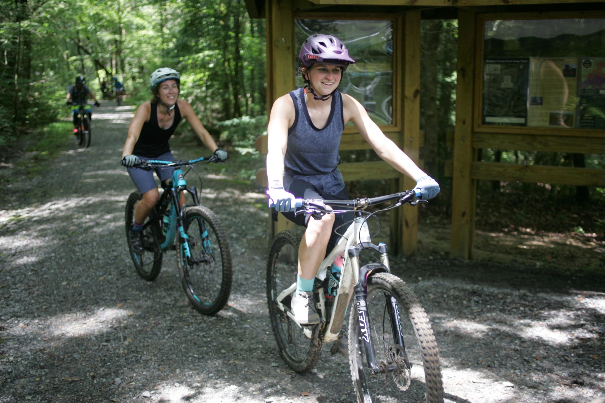 Two women are riding mountain bikes along a gravel trail surrounded by lush green trees. One woman is in the foreground, smiling as she pedals, wearing a black tank top and a purple helmet. The second woman is visible in the background, also riding a bike. A wooden information sign can be seen along the trail. Weed Patch Mountain Trail mountain bike trail.