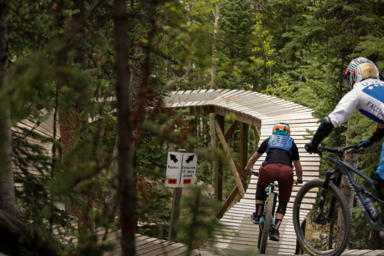 Two mountain bikers navigate a wooden elevated trail winding through a lush forest. A directional sign indicates paths for "Bypass" and "Corkscrew." The trail is surrounded by trees, creating a natural setting for an adventurous ride.