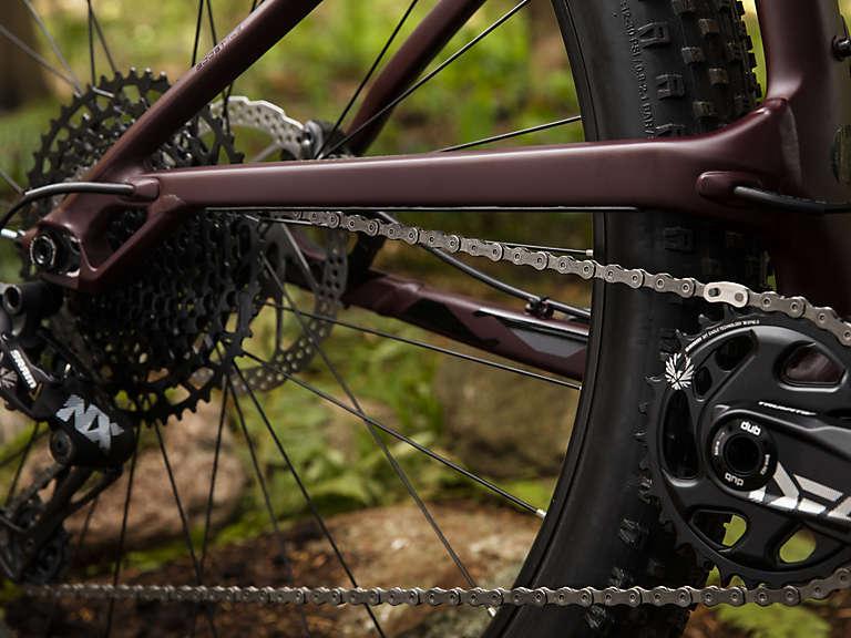 Trek Stache 7: Close-up view of a mountain bike's rear drivetrain, featuring a durable chain, cassette, and crankset. The bike is positioned on a natural background with greenery and rocks. The frame is a rich burgundy color, highlighting its sleek design and components.