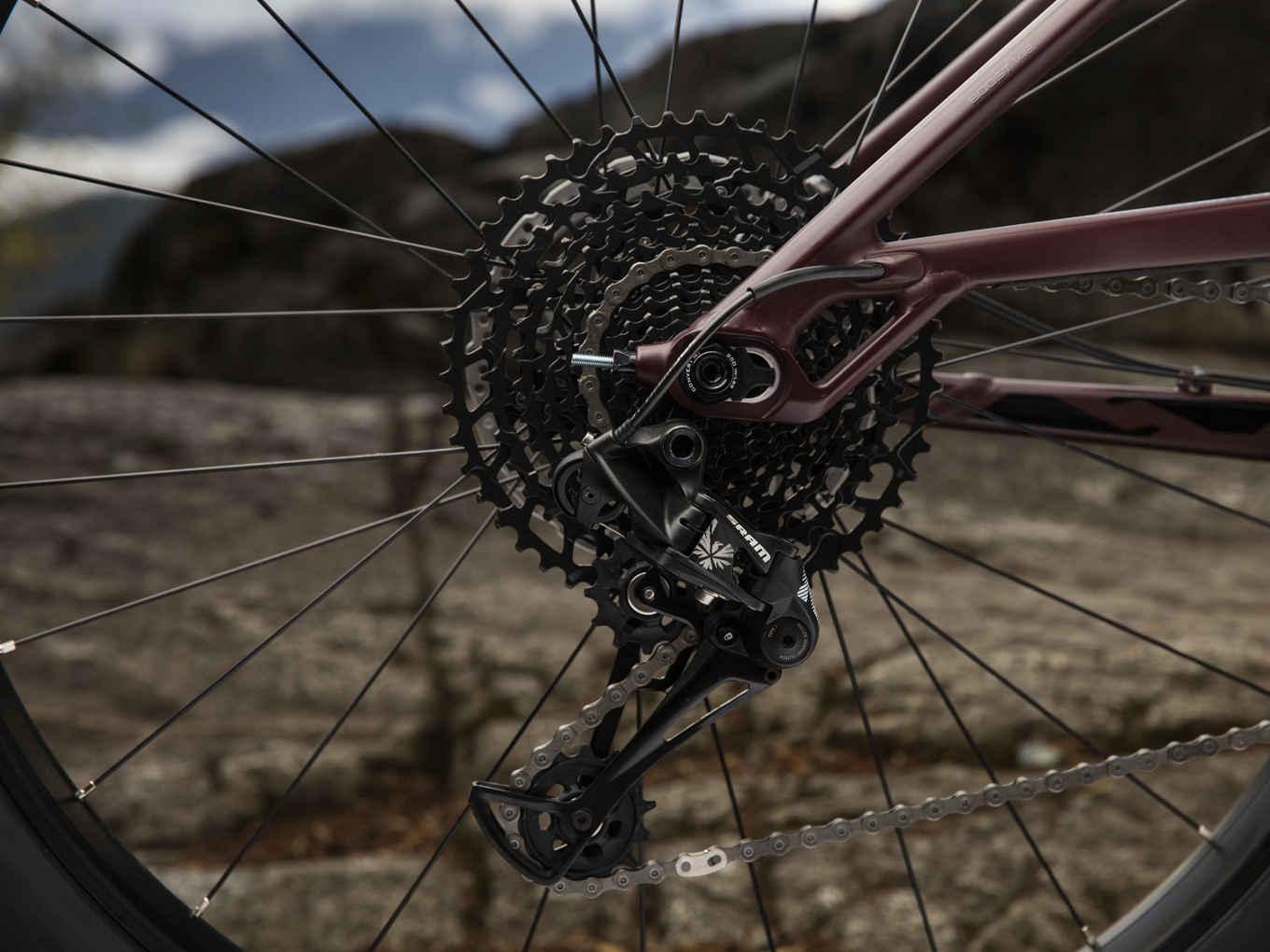 Trek Stache 7: Close-up view of a bicycle rear derailleur and cassette, showcasing intricate components and chain details against a rocky outdoor background. The image highlights the mechanical precision of the bike's drivetrain system.