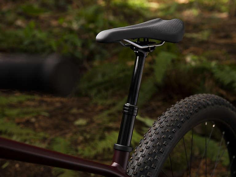 Trek Stache 7: Close-up view of a bicycle saddle and seat post amidst a forest background, featuring a textured black saddle and a sleek black post, with a blurred bicycle wheel partially visible.
