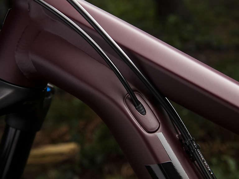 Trek Stache 7: Close-up view of the frame of a bicycle, showcasing a sleek, dark maroon finish and the integrated cables running smoothly along the frame. The background features blurred greenery, highlighting the bicycle's design and craftsmanship.