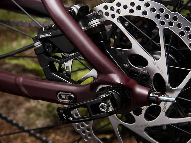 Trek Stache 7: Close-up of a bicycle's rear brake assembly, featuring a maroon frame component, disc brake, and chainstay. The image showcases detailed mechanical elements, including the Shimano brake and rotor, against a blurred natural background.