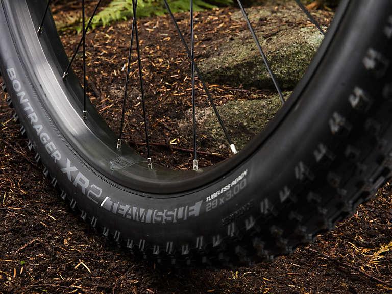 Trek Stache 7: Close-up of a mountain bike tire resting on dirt and gravel, featuring a Bontrager XR2 Team Issue tire with the specifications clearly visible: "29x2.30" and "tubeless ready." The tire is mounted on a dark rim, with spokes partially visible, set against a natural background with ferns and stones.