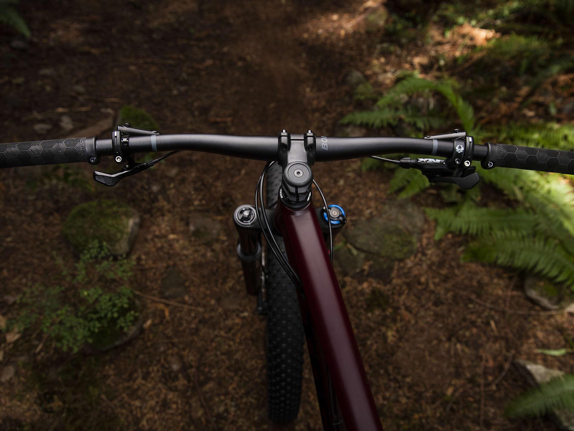 Trek Stache 7: Alt text: A close-up view of a mountain bike's handlebars, featuring textured grips, gear shifters, and bicycle controls, set against a natural background of dirt, rocks, and green ferns.
