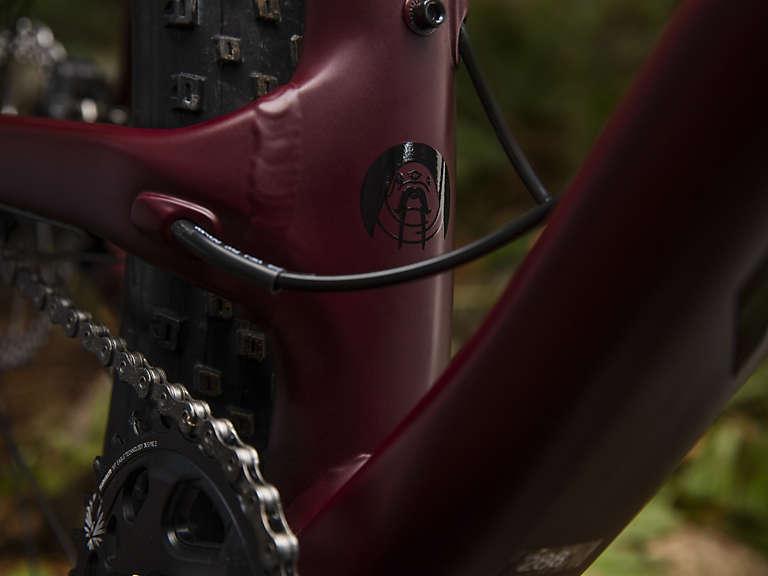 Trek Stache 7: A close-up view of a maroon bicycle frame featuring a distinctive logo, with visible cable routing and a dark tire in the background. The image captures the detailed craftsmanship of the bike's design.