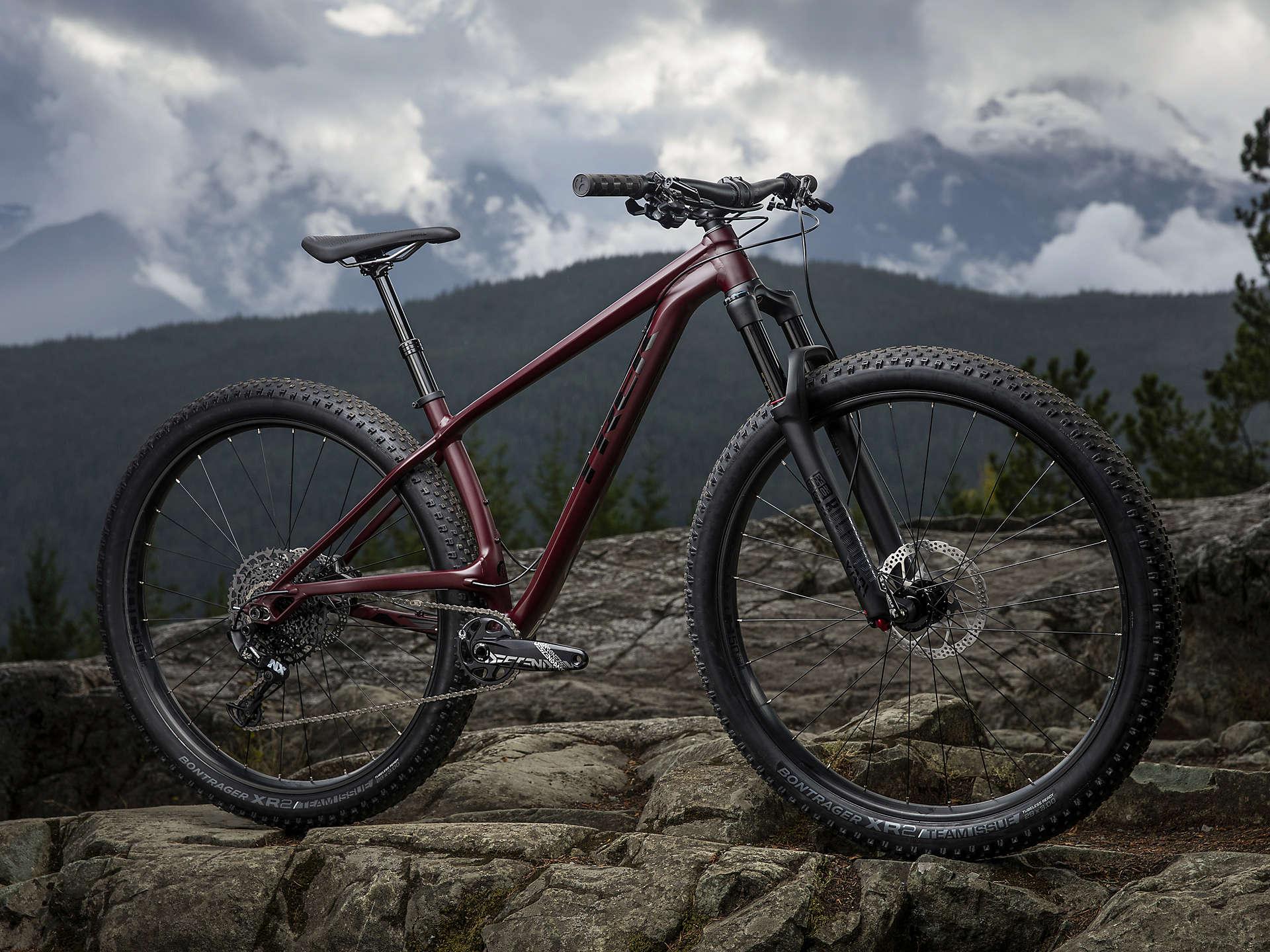 Trek Stache 7: A mountain bike with a sleek maroon frame is positioned on rocky terrain, showcasing sturdy tires and suspension. The backdrop features a dramatic sky with clouds and distant mountains, emphasizing an outdoor adventure setting.