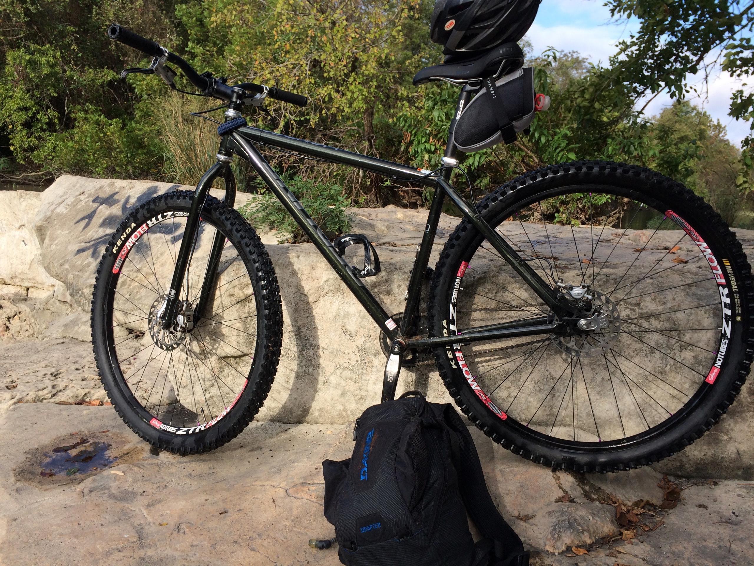 Redline Monocog 29er: A black mountain bike with thick tires stands on a rocky surface, accompanied by a black backpack. The bike features a disc brake and a helmet attached to the seat, surrounded by greenery and trees in the background.