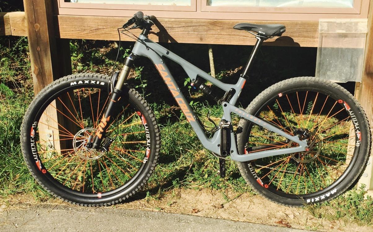Santa Cruz Santa Cruz Tallboy CC: A modern mountain bike with a gray frame and orange accents is parked next to a wooden signpost. The bike features wide tires and a front suspension fork, set against a backdrop of grass and a paved path.