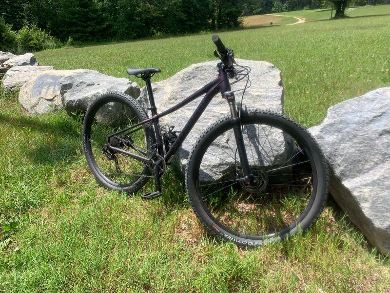 A mountain bike resting on a grassy area near large rocks, with trees and a path visible in the background. The bike has a dark frame and rugged tires, suggesting it is designed for off-road use. DuPont State Recreational Forest mountain bike trail.