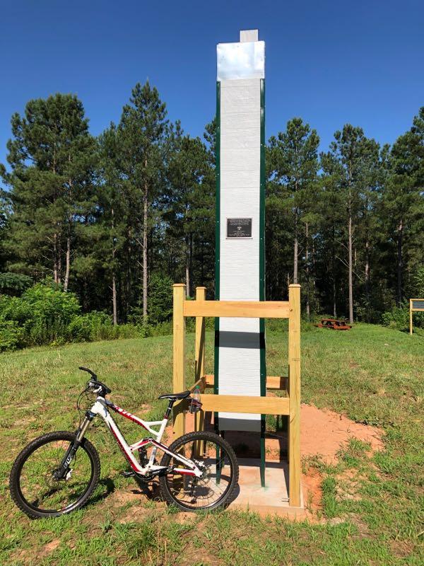 A white mountain bike stands next to a tall, white monument with a plaque, surrounded by lush green grass and pine trees. The monument is secured with wooden supports, and there are picnic tables visible in the background under a clear blue sky. Talking Rock Nature Preserve mountain bike trail.