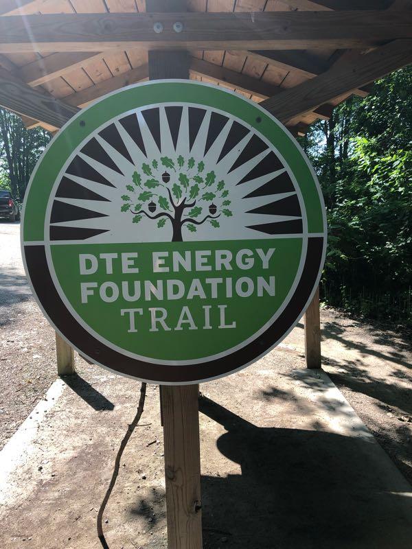 Sign for the DTE Energy Foundation Trail, featuring a circular design with a tree and acorns, set against a green background. The sign is mounted on a wooden post under a shelter, indicating the beginning of the trail. Surrounding vegetation is visible in the background. DTE Energy Foundation Trail mountain bike trail.