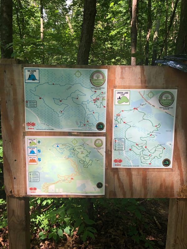 Trail maps displayed on a wooden board in a wooded area, featuring routes for Big Lake, Green Lake, and another loop. Each map includes marked trails, icons indicating various amenities, and outlines of the lakes. The surrounding environment is lush with greenery, typical of a forested setting. DTE Energy Foundation Trail mountain bike trail.
