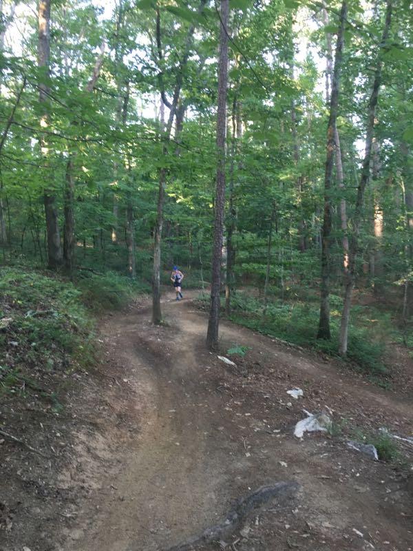 A person running along a winding dirt trail in a dense, green forest with tall trees and sunlight filtering through the leaves. Dwelling Loop mountain bike trail.