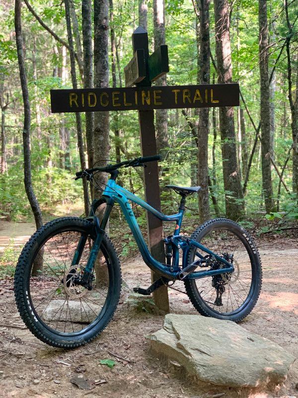 A mountain bike parked next to a wooden trail sign that reads "RIDGELINE TRAIL," surrounded by a lush green forest with tall trees and a dirt path leading into the woods. Ridgeline Trail #65 mountain bike trail.