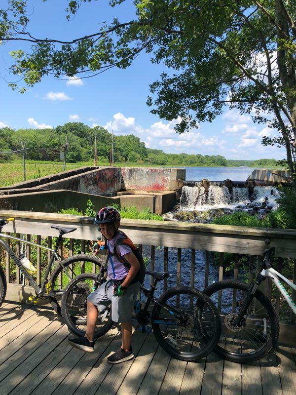 A child wearing a helmet and casual clothing sits on a wooden deck beside several bicycles, with a flowing waterfall and lush greenery in the background under a clear blue sky. Ryan park mountain bike trail.