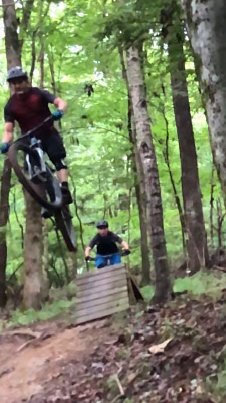 Two mountain bikers riding through a wooded trail. The first rider is airborne, performing a jump over a wooden ramp, while the second rider is positioned on the ground, preparing to follow. The lush greenery of the forest surrounds them. Lock 4 mountain bike trail.