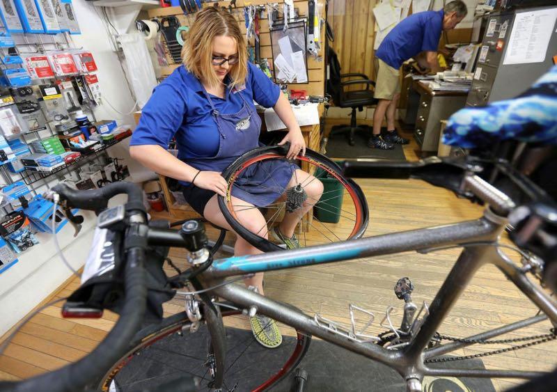 A woman is repairing a bicycle tire in a bike shop, sitting on a stool with a tire in her hands. She is wearing a blue shirt and overalls, focused on her work. In the background, a man is organizing items at a counter, surrounded by various bicycle tools and parts displayed on the wall. The shop has a wooden floor and a relaxed, busy atmosphere.