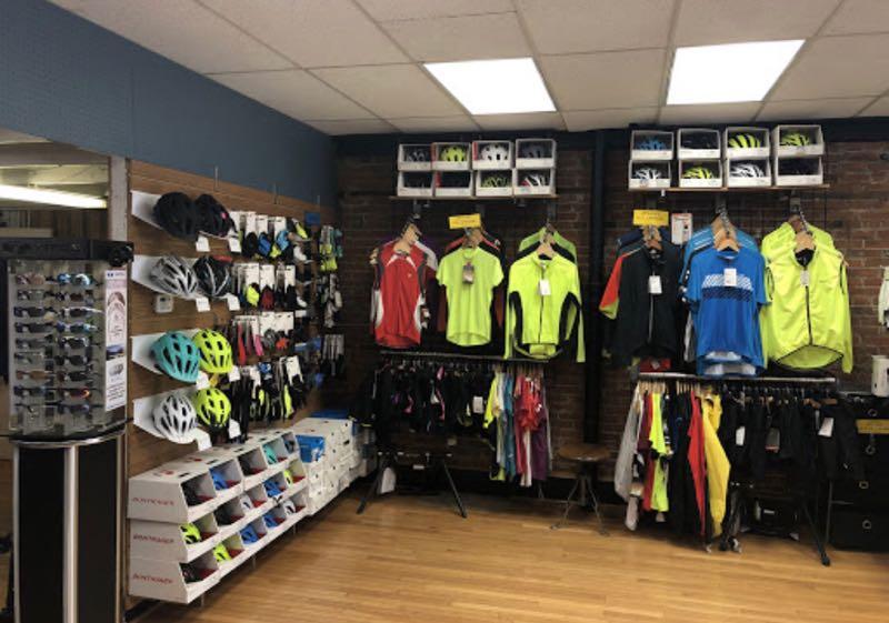 A brightly lit retail store showcasing cycling gear. The left wall features a display of various helmets and sunglasses, while the right side has a colorful assortment of cycling jerseys and jackets hanging on racks. The floor is wooden, and shelves at the bottom hold additional cycling shoes and accessories.