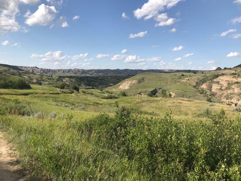 A scenic view of rolling hills and grassy fields under a partly cloudy sky. The landscape features green vegetation, gentle slopes, and distant rocky formations, creating a tranquil natural setting. Long X mountain bike trail.