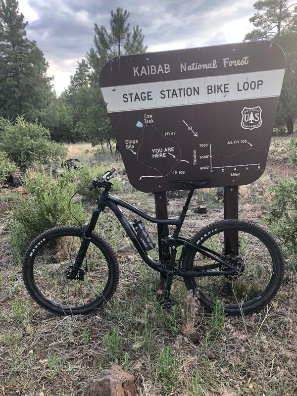 A mountain bike parked next to a sign for the Stage Station Bike Loop in Kaibab National Forest, surrounded by greenery and pine trees, with a cloudy sky overhead. The sign features a map indicating the bike trail and shows the location labeled "YOU ARE HERE." Stage Station Loop mountain bike trail.