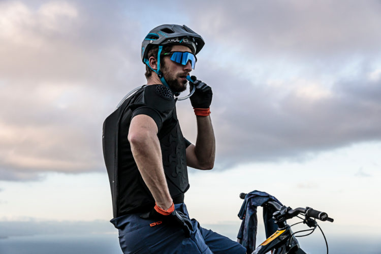 A mountain biker wearing a helmet and sunglasses pauses to take a drink from a hydration tube, overlooking a scenic landscape with clouds in the background. He is dressed in a black shirt and blue shorts, with protective gear visible on his shoulders. A bike is positioned nearby.