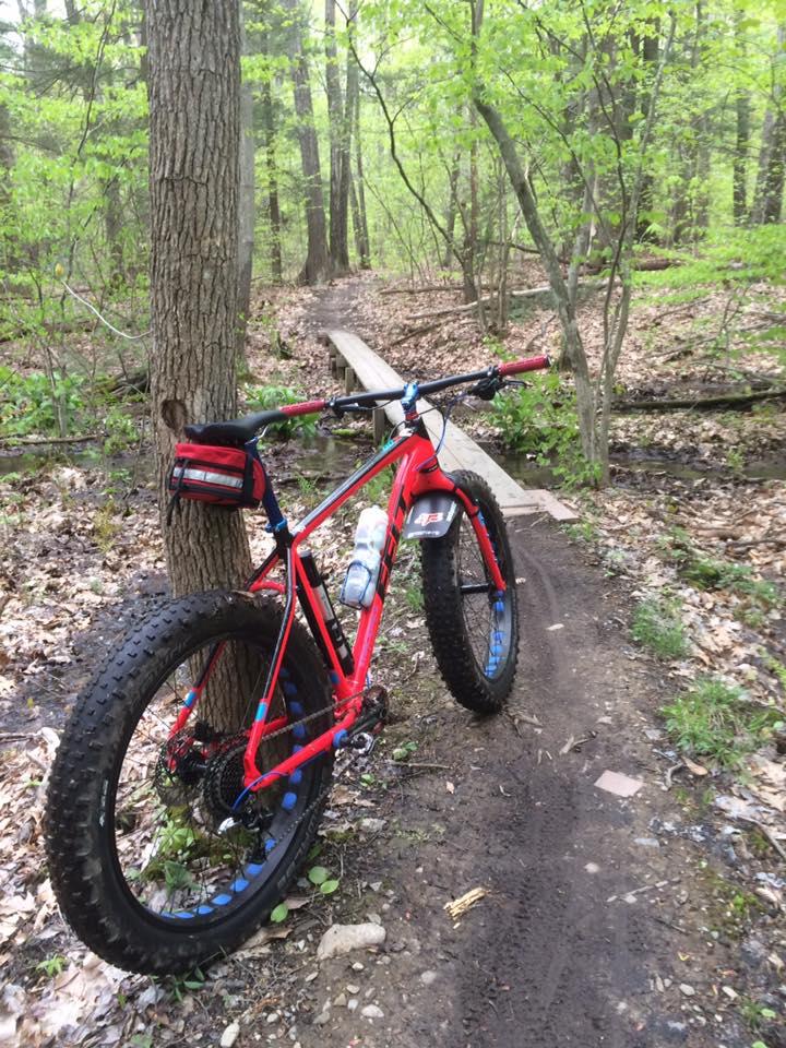 A red fat tire bike leaning against a tree on a dirt trail in a lush green forest, with a wooden bridge in the background and a water bottle attached to the bike frame. Stratham Hill Park mountain bike trail.