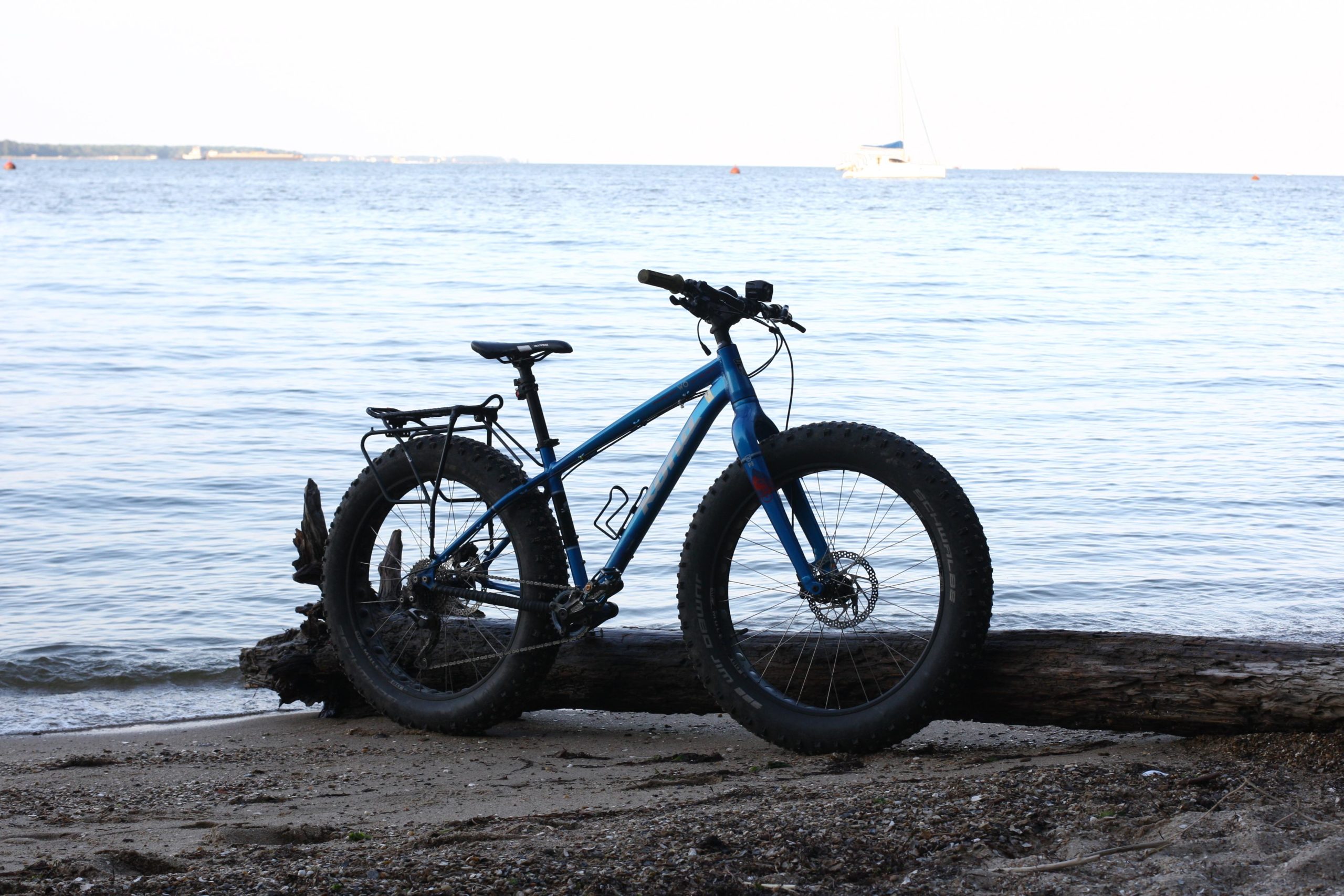 A blue fat bike resting on a log at the beach, with calm water and a distant sailboat in the background. The scene captures a serene coastal landscape, showcasing the bike in a recreational setting. York River State Park mountain bike trail.