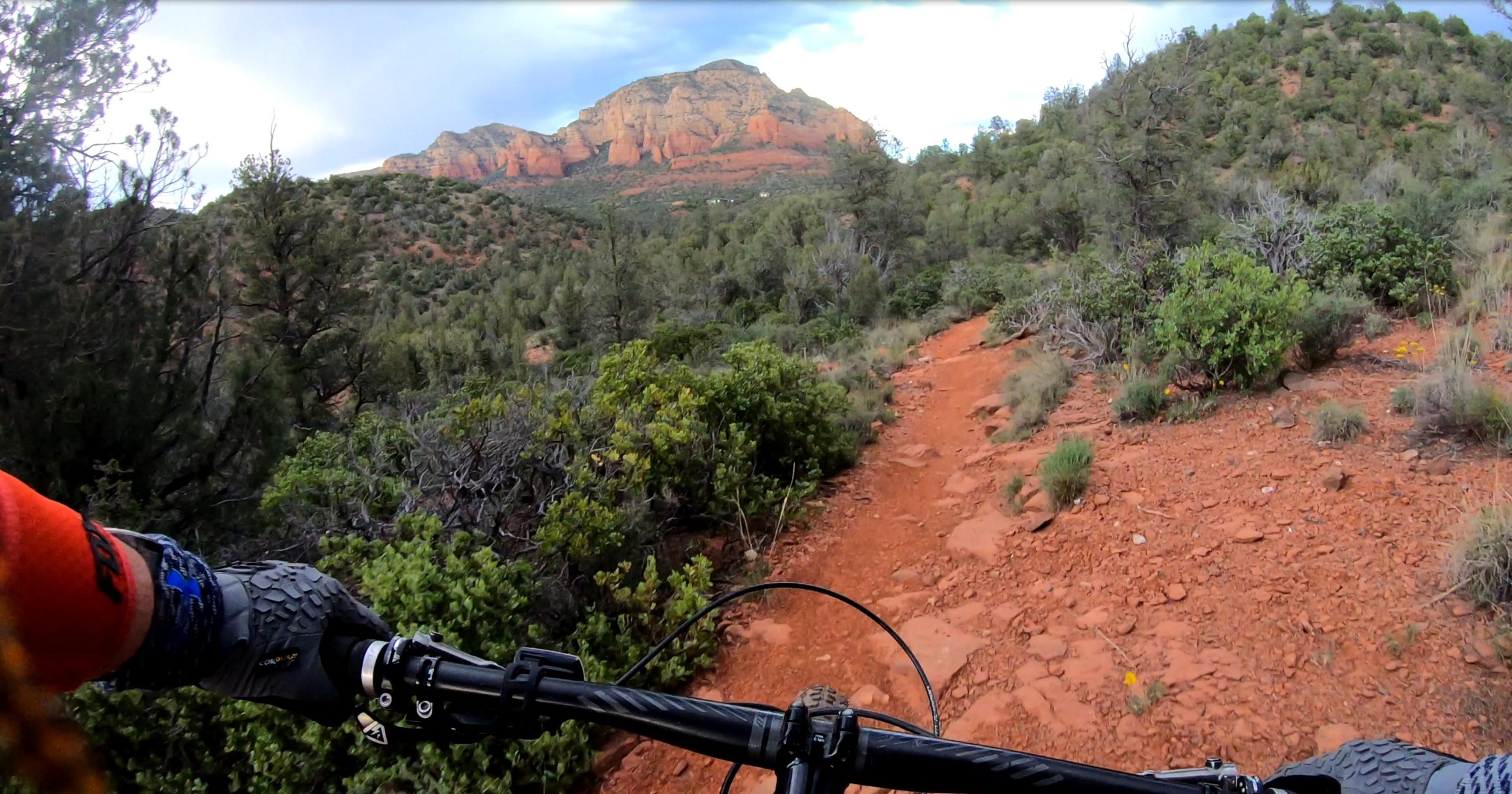 A mountain biker's view from the handlebars, depicting a rugged, winding trail through green shrubs and trees, with red rock formations rising in the background under a partly cloudy sky. Bell Rock Pathway mountain bike trail.