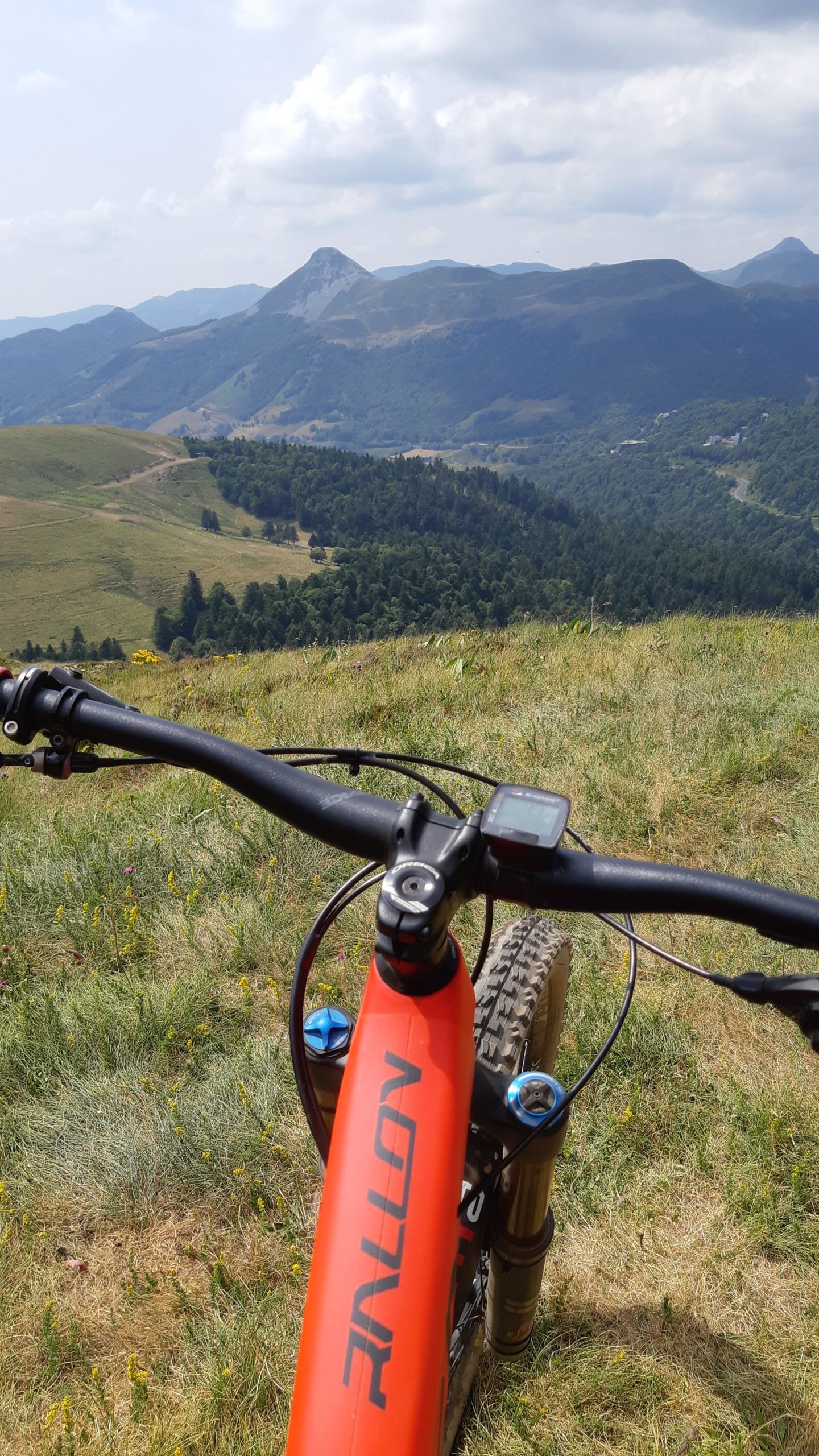 A mountain bike is positioned on a grassy hillside, with a panoramic view of rolling mountains and valleys in the background. The bike