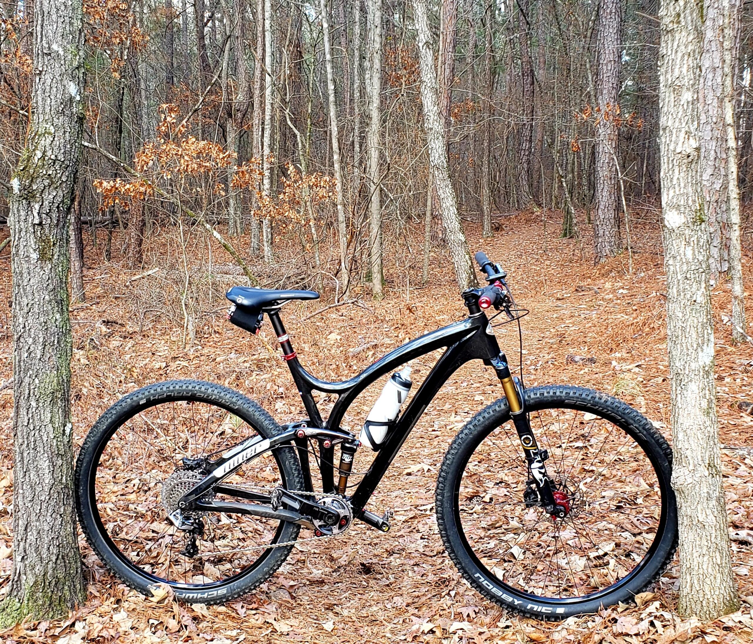 Niner Jet 9 RDO: A black mountain bike leaned against a tree in a wooded area, surrounded by fallen leaves and tall trees. The forest path is visible in the background, indicating a natural outdoor setting suitable for biking.