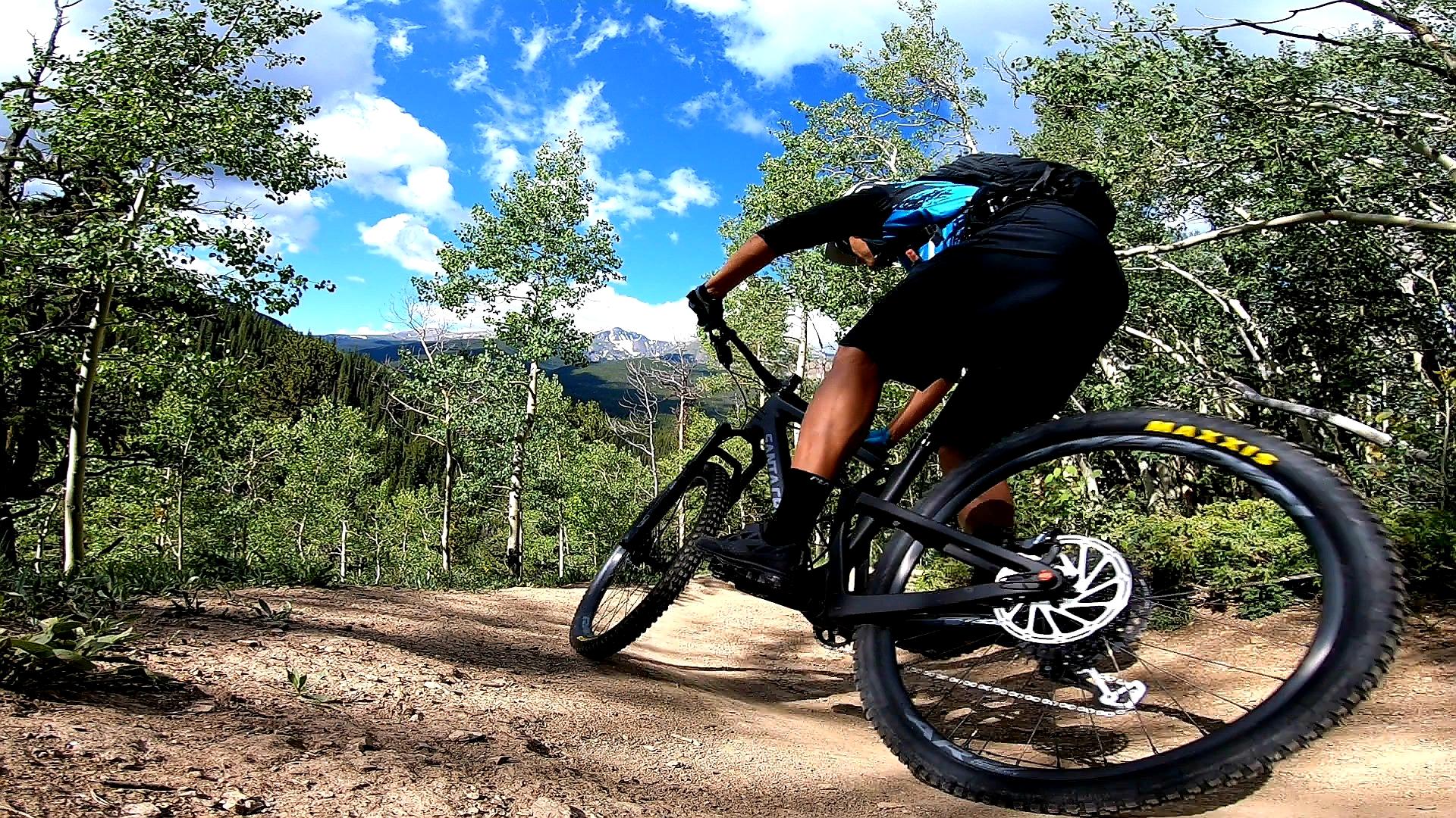 A mountain biker navigating a dirt trail surrounded by greenery and mountains, leaning into a turn while riding a black mountain bike, against a backdrop of blue skies with fluffy clouds. Aspen Alley mountain bike trail.