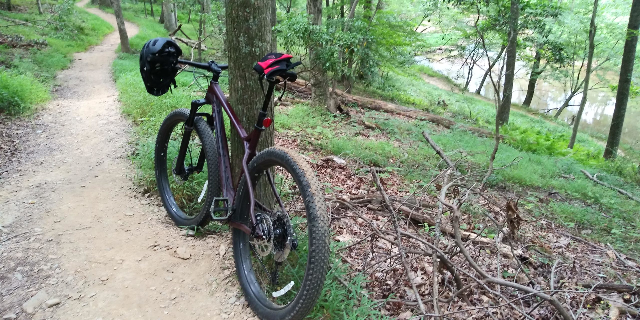 A mountain bike parked on a dirt trail surrounded by trees and greenery, with a helmet resting on the handlebars. A river can be seen in the background, indicating a scenic outdoor biking location. Seneca Creek Greenway Trail mountain bike trail.