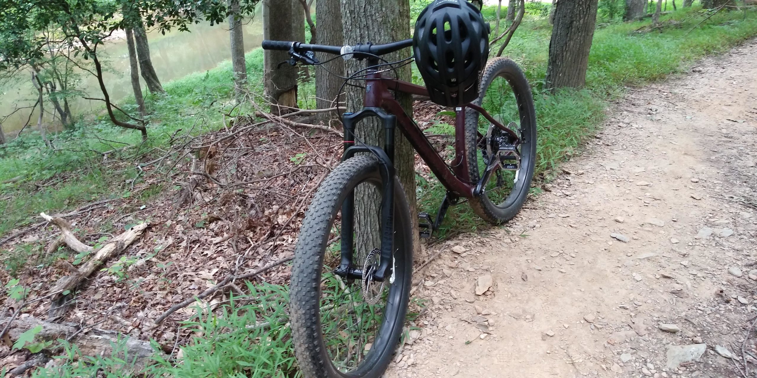A mountain bike with a black helmet resting on the handlebars, positioned next to a tree along a dirt path in a lush green forested area. A calm body of water is visible in the background, partially obscured by trees and foliage. Seneca Creek Greenway Trail mountain bike trail.