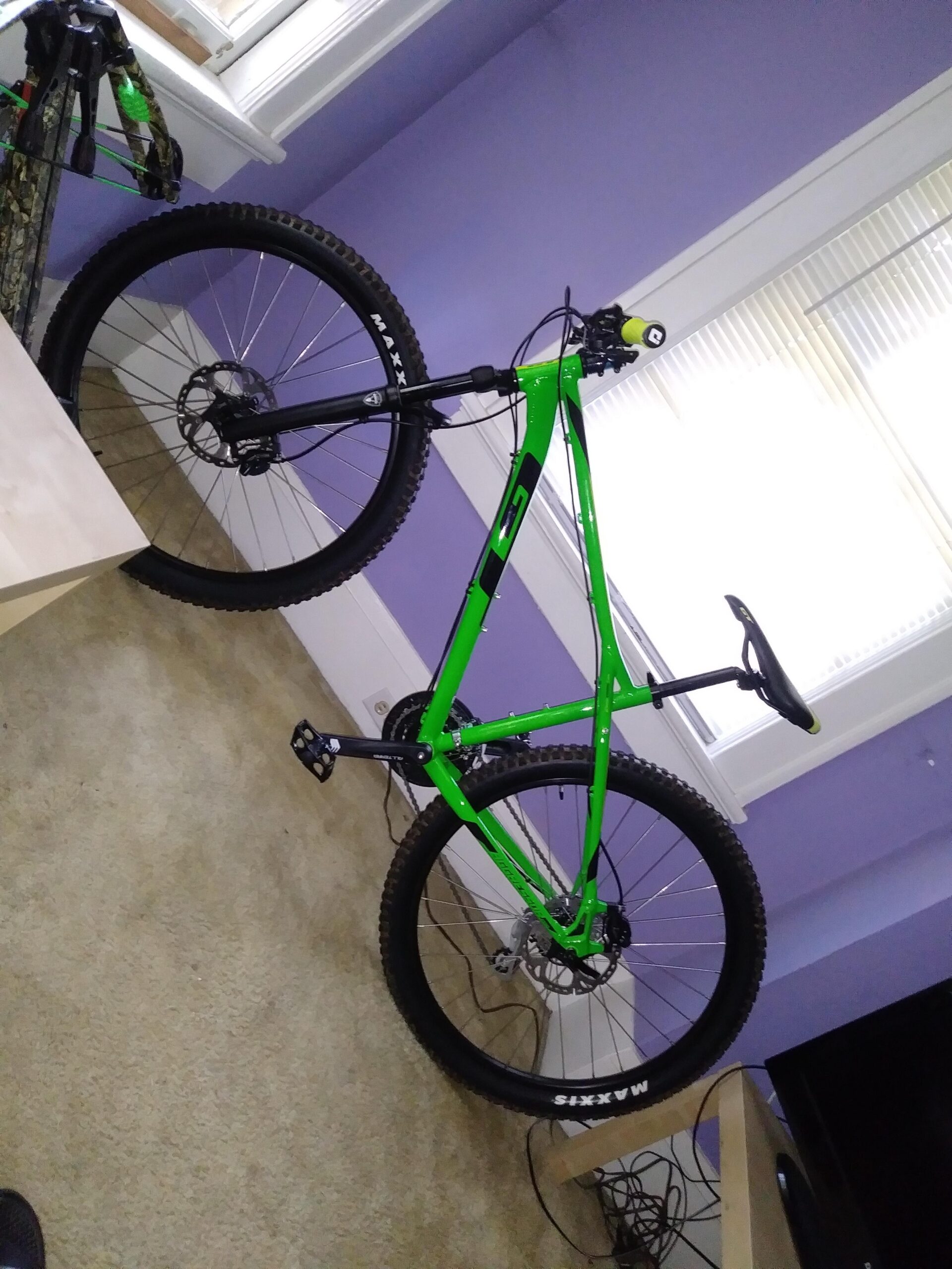 A bright green mountain bike displayed indoors, leaning against a wall with a purple-colored backdrop. The bike features thick tires, sturdy frame, and a black seat. Nearby, another bike is partially visible, along with a beige table and some electrical cords on the carpeted floor.