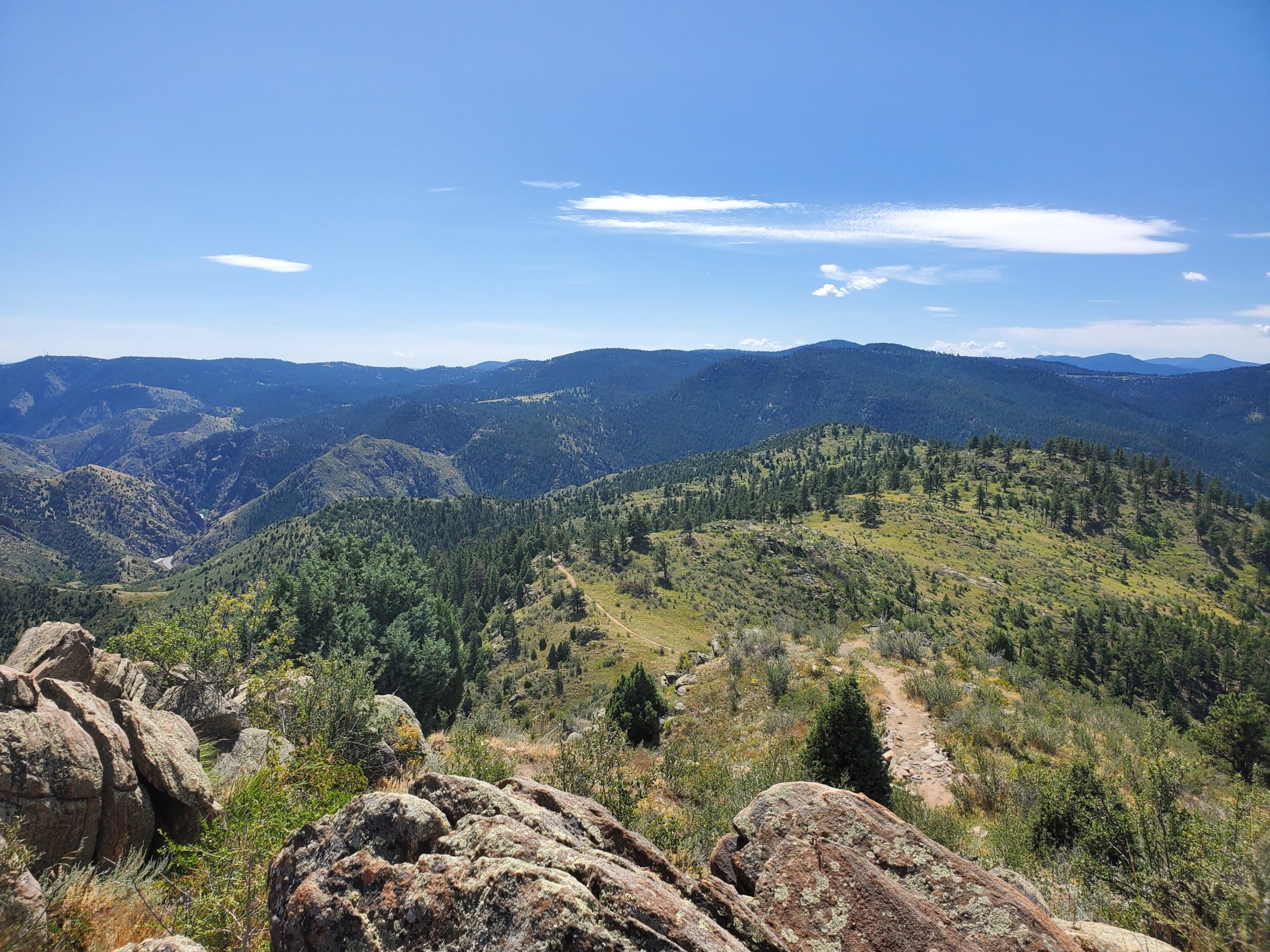 A panoramic view of rolling hills and mountains under a clear blue sky, with rocky formations in the foreground. The landscape features lush green trees and vibrant vegetation, showcasing a serene natural environment. Centennial Cone Park mountain bike trail.