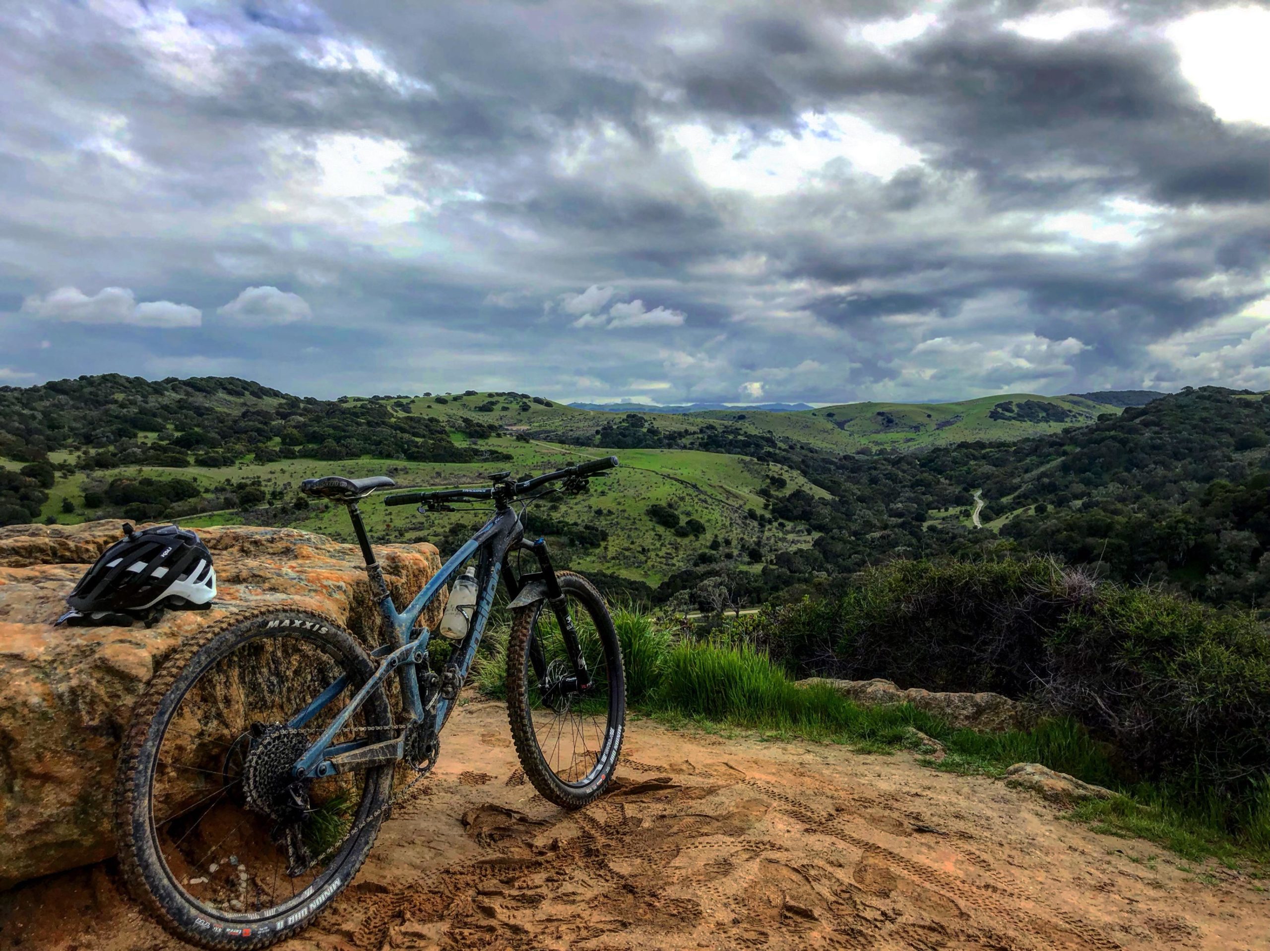 A mountain bike resting on a rock outcrop, with a white bicycle helmet nearby. The background features lush green hills under a cloudy sky, creating a scenic nature landscape. Fort Ord Public Lands mountain bike trail.