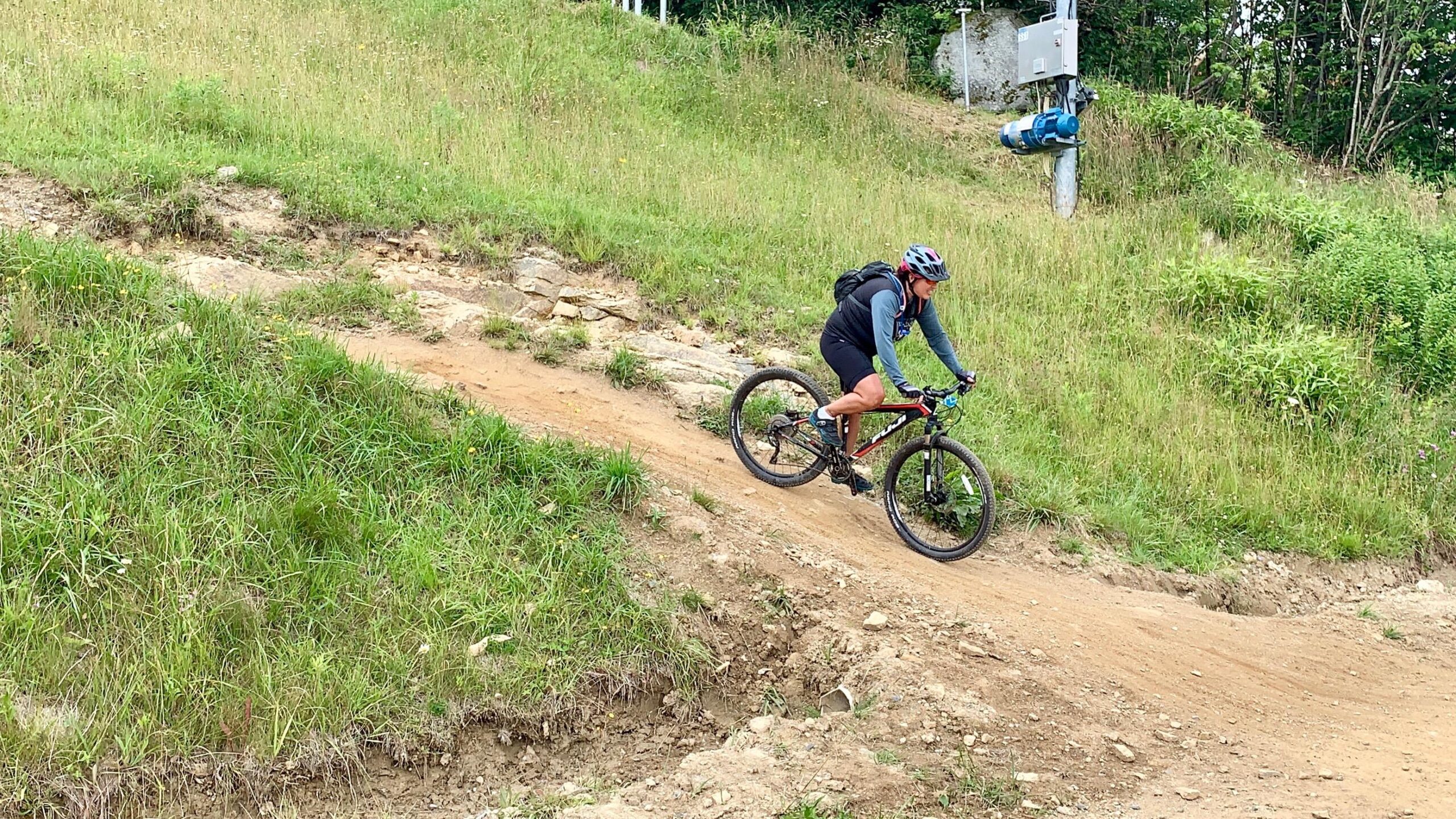 Fuji Tahoe 1.5: A mountain biker navigating a dirt trail on a grassy hillside, surrounded by vibrant greenery. The cyclist is wearing a helmet and a backpack, focusing on the challenging terrain ahead.