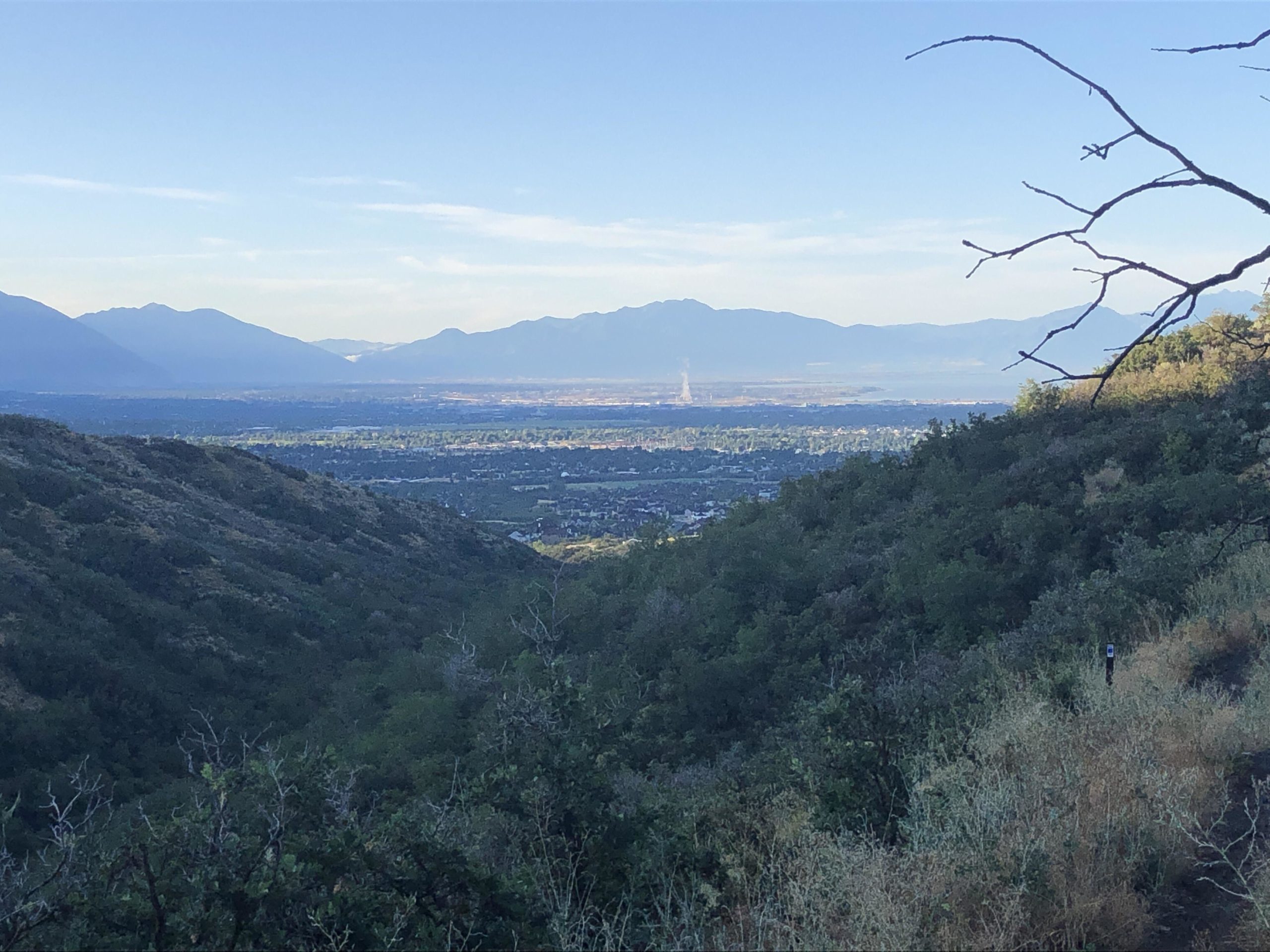 A panoramic view of a valley surrounded by mountains, featuring lush green vegetation on the slopes and a clear blue sky. The landscape shows a distant city and a hint of industrial activity with wisps of smoke visible in the far background. Achtung Baby mountain bike trail.