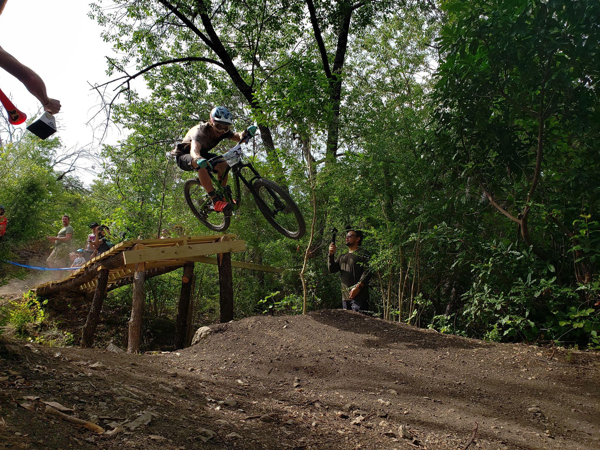 A mountain biker performing a jump over a wooden ramp in a forested area, with spectators watching nearby. The biker is wearing a helmet and protective gear, surrounded by greenery and dirt paths. Sansom Park mountain bike trail.