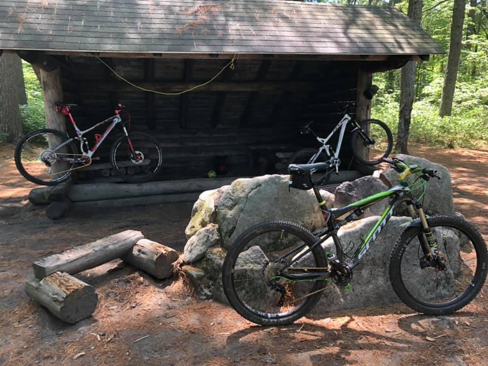 A shaded resting area in a wooded setting, featuring two mountain bikes parked on a large rock and a log. The structure has a wooden roof and a backdrop of trees. Sunlight filters through the foliage, creating a serene outdoor atmosphere. Bear Brook mountain bike trail.