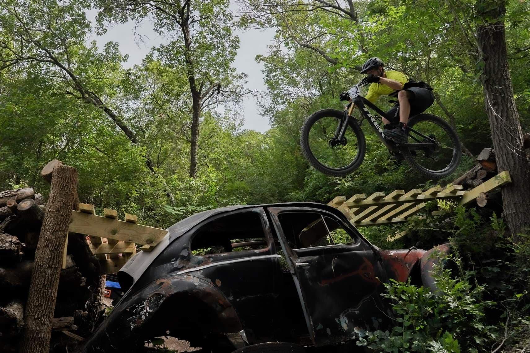 A mountain biker performs a jump over a wooden ramp supported by logs, with an old, partially obscured black car in the foreground. Lush green foliage surrounds the scene, indicating a natural outdoor setting. Sansom Park mountain bike trail.