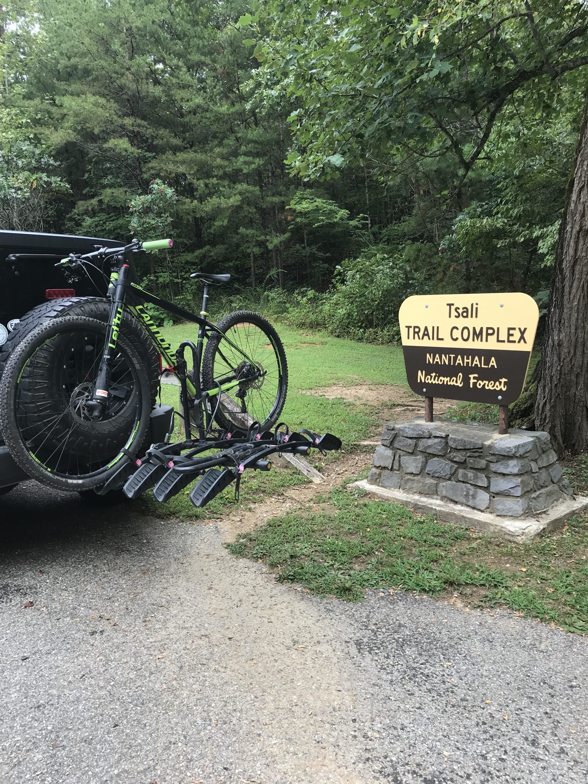 A mountain bike mounted on the back of a vehicle next to a sign for the Tsali Trail Complex in Nantahala National Forest, surrounded by lush green trees and grassy areas. The scene captures a peaceful outdoor setting ideal for biking and nature activities. Tsali Recreation Area mountain bike trail.
