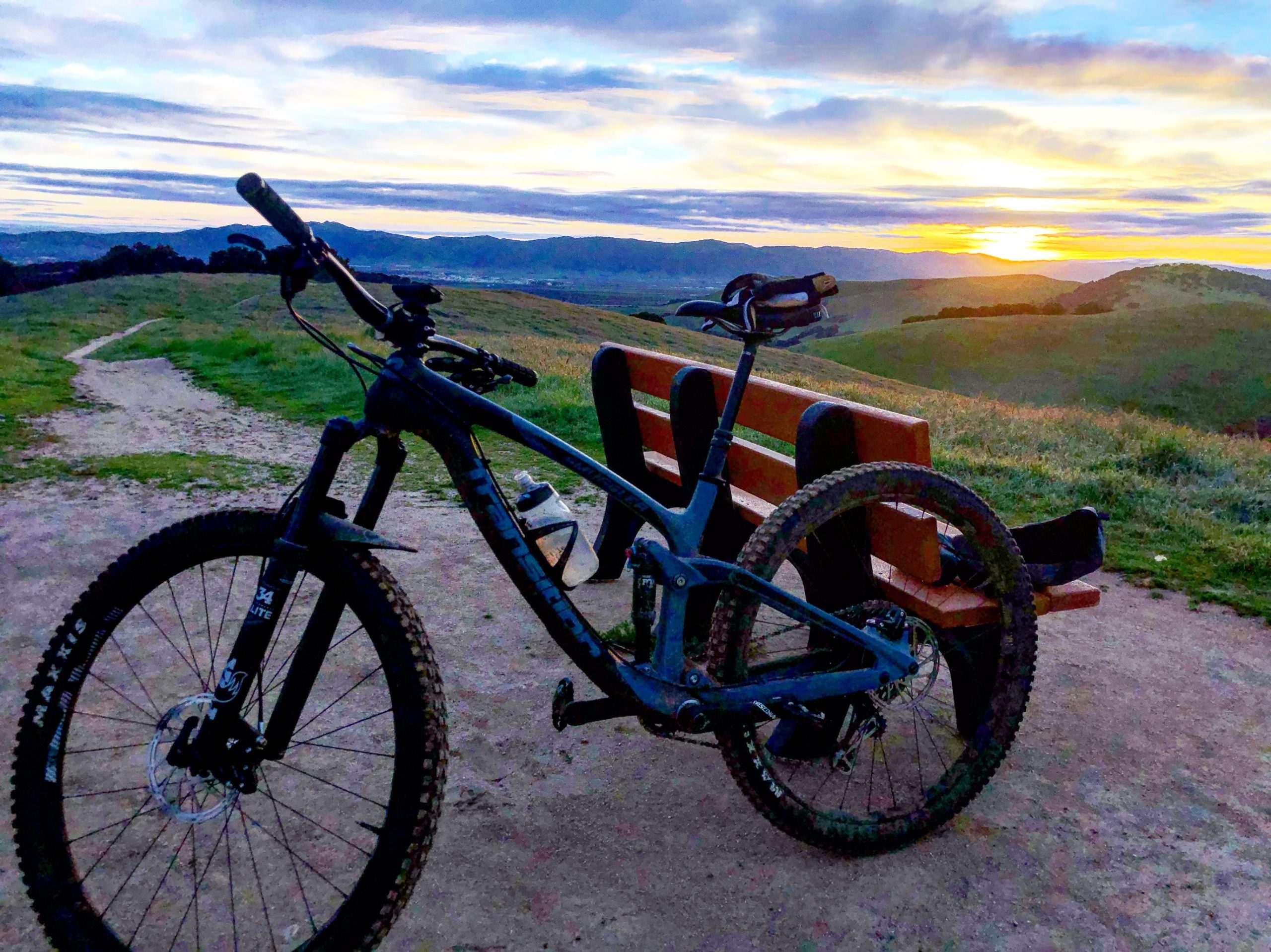 A mountain bike parked next to a wooden bench on a grassy hillside, with a vibrant sunset casting warm colors across the sky and illuminating distant mountains. A dirt path winds through the landscape, inviting exploration. Fort Ord Public Lands mountain bike trail.