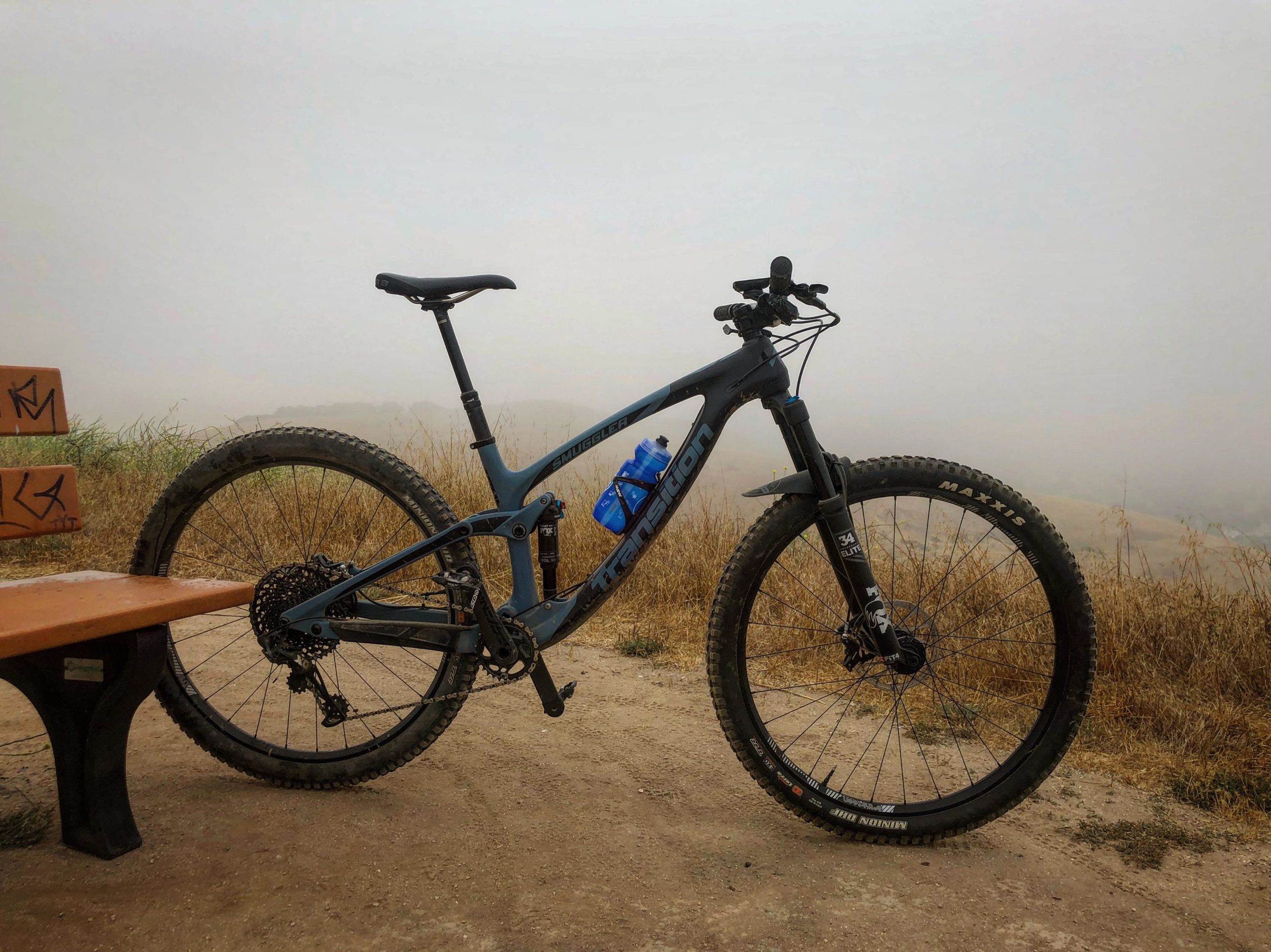 A mountain bike rests on a gravel path near a bench, with tall grasses and a foggy background. The bike features a blue and black design, a water bottle mounted on the frame, and large knobby tires, suggesting readiness for rugged terrain. Fort Ord Public Lands mountain bike trail.