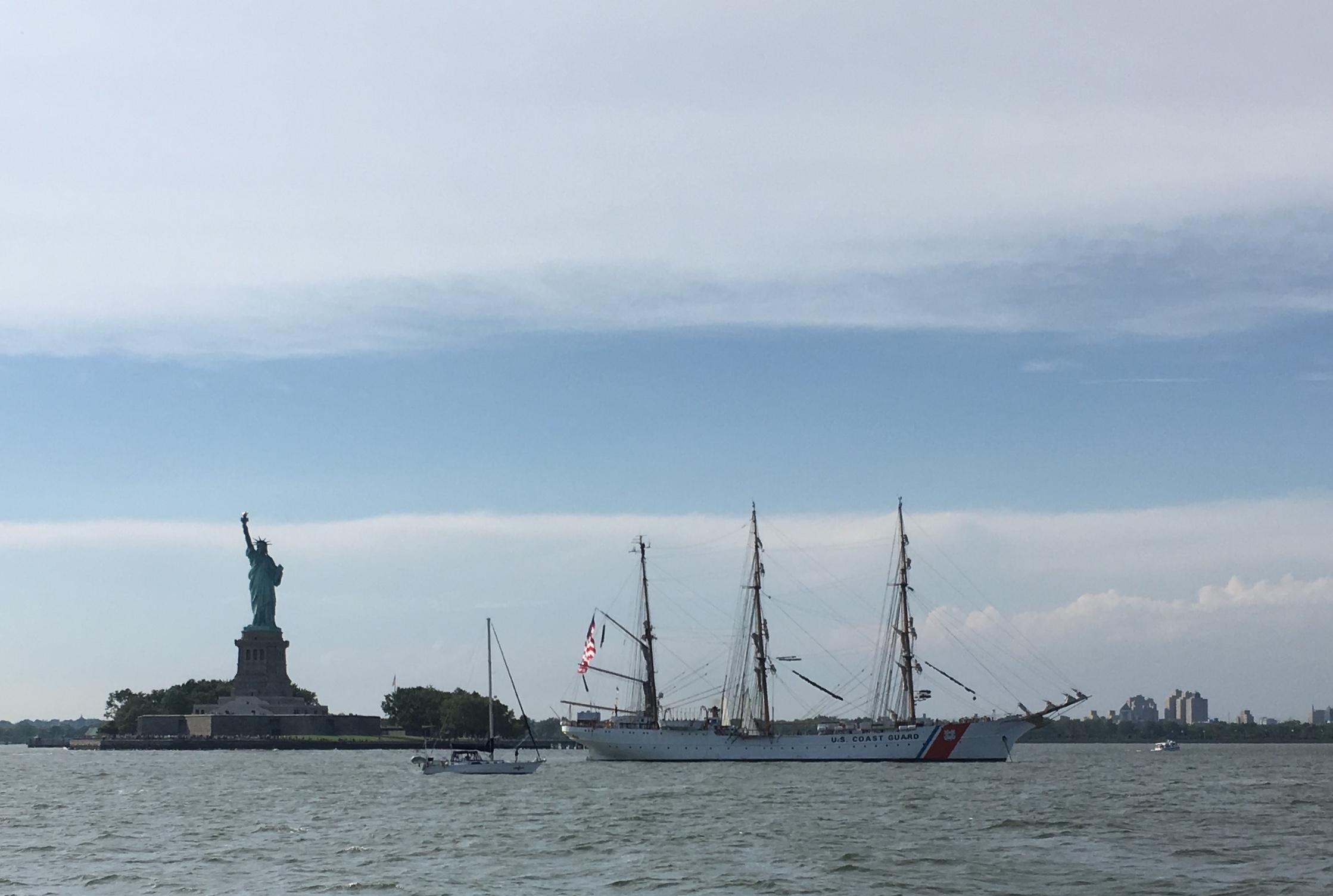 A scenic view of the Statue of Liberty on Liberty Island, with a tall sailing ship marked "U.S. Coast Guard" navigating the water in the foreground. A smaller sailboat is also visible nearby, set against a backdrop of a clear blue sky with scattered clouds. West Street Greenway mountain bike trail.