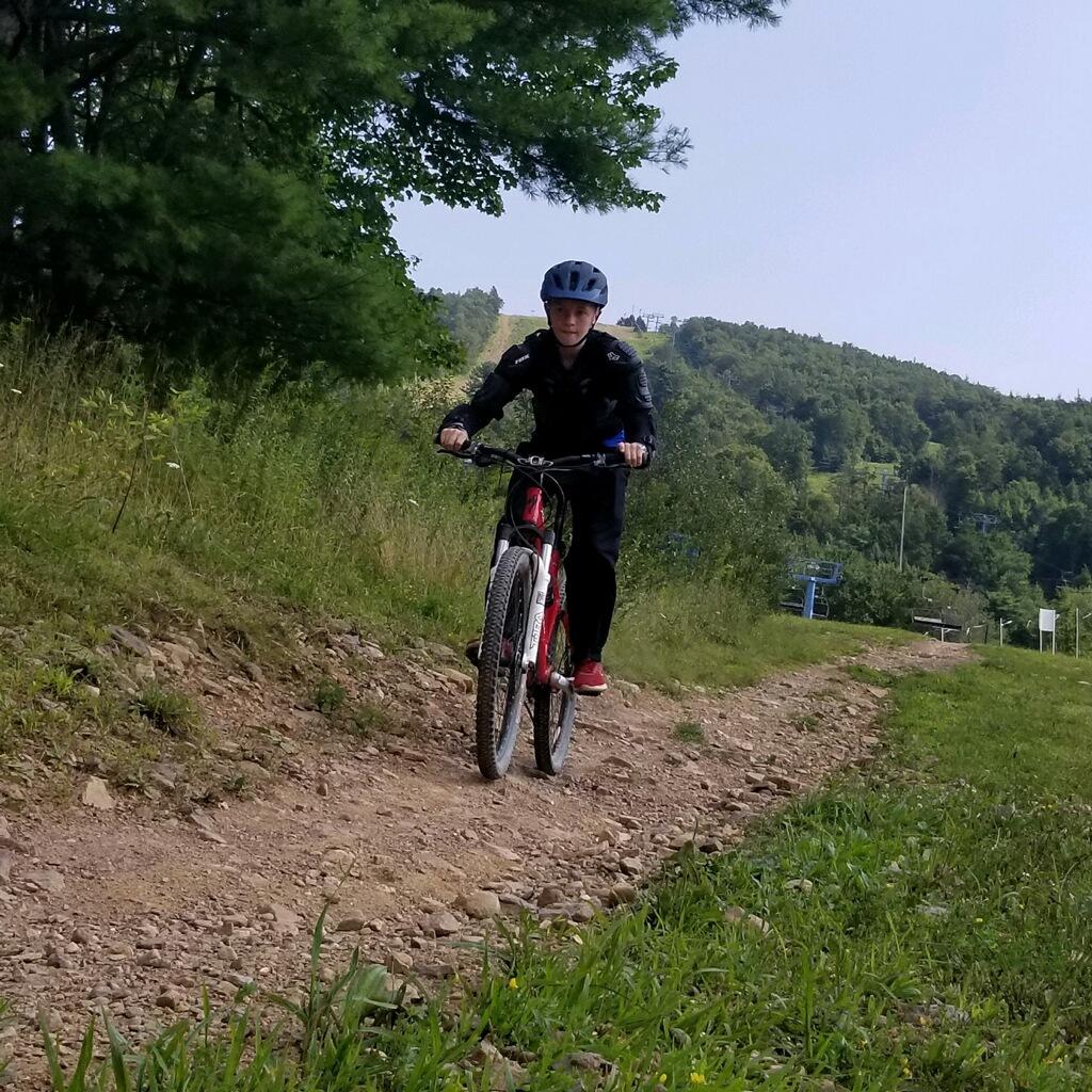 Specialized Specialized XC Comp MTB: A young cyclist riding a mountain bike on a dirt path surrounded by greenery and trees. The cyclist is wearing a helmet and a black jacket, focused on the trail ahead, with a hillside visible in the background.